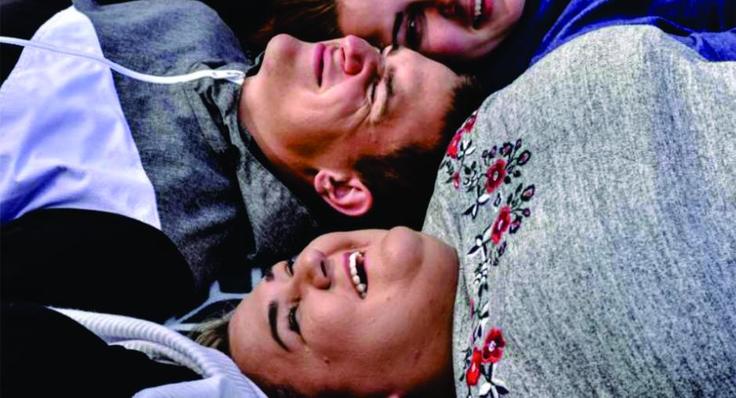 Three people lying close together, smiling and laughing, wearing sweaters with varying patterns.