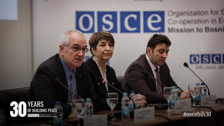 Three people seated at a conference table with OSCE logos in the background.