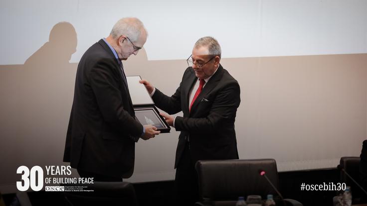 Two men in suits exchange a plaque at an event, seated audience in foreground, with a "30 Years" graphic on the corner.