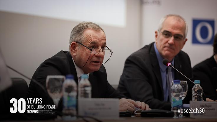 Two men in suits speaking at a conference table with microphones and water bottles.