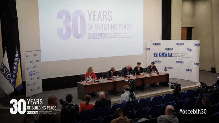 Panel discussion in a conference room with four speakers seated at a table, audience watching. 30 Years of Building Peace event backdrop.