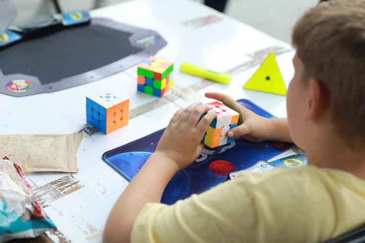 A child in a yellow shirt solves a Rubik's Cube on a table with other puzzles.