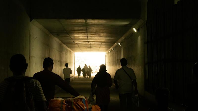 Silhouettes of people walking through a dimly lit tunnel toward bright light, some carrying bags.