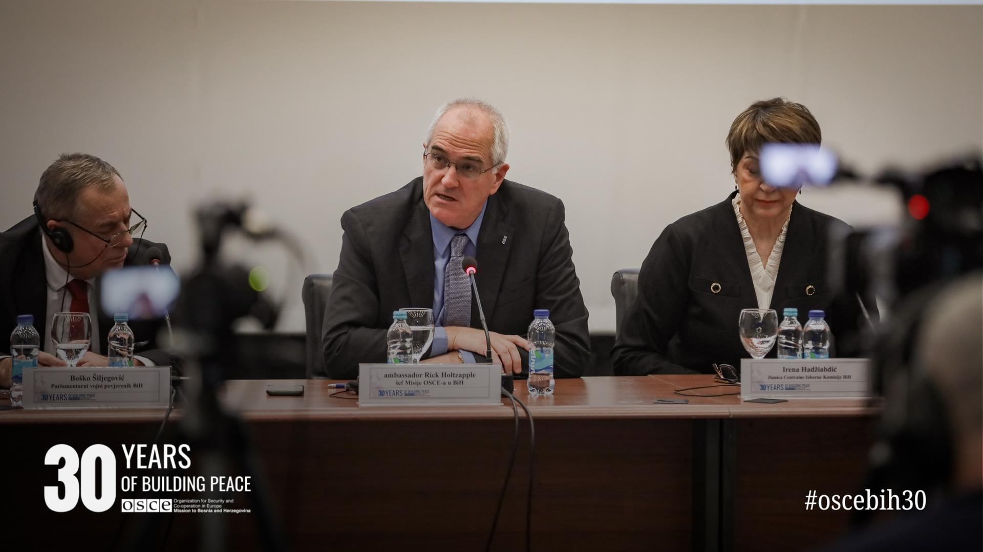 Three people sitting at a conference table with microphones and papers, engaged in discussion.