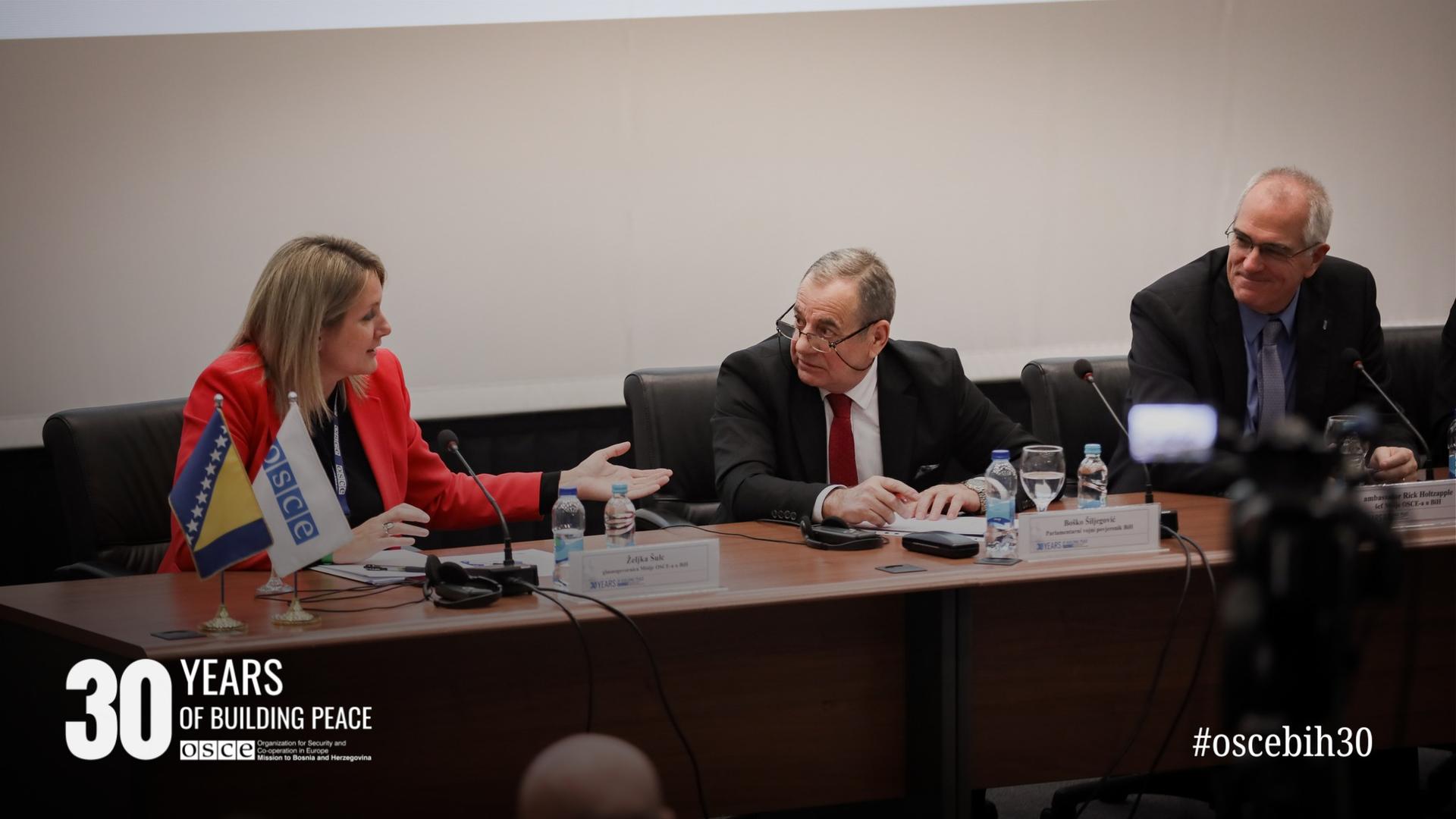 Three people in a panel discussion; a woman gesturing next to two men in suits, flags on the table.