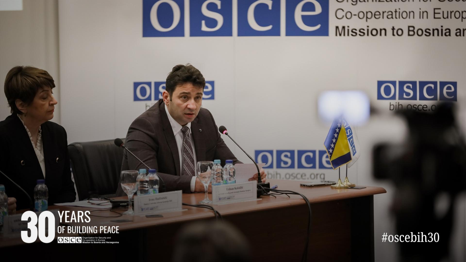 A man speaks at a conference table with OSCE branding, accompanied by two others. A Bosnian flag and camera are visible.